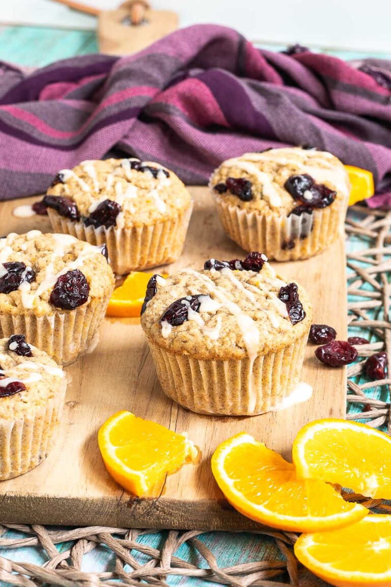 Orange cranberry muffins drizzled with a white glaze on a wooden board surrounded by lots of dried cranberries and fresh orange slices.