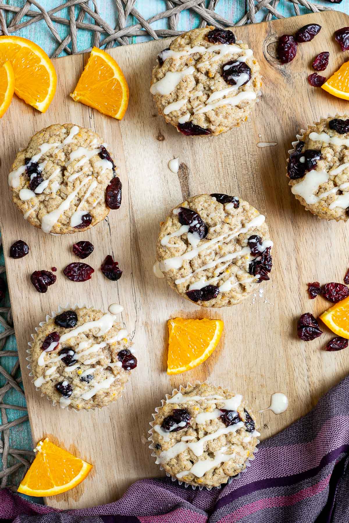 Orange cranberry muffins drizzled with a white glaze on a wooden board surrounded by lots of dried cranberries and fresh orange slices.