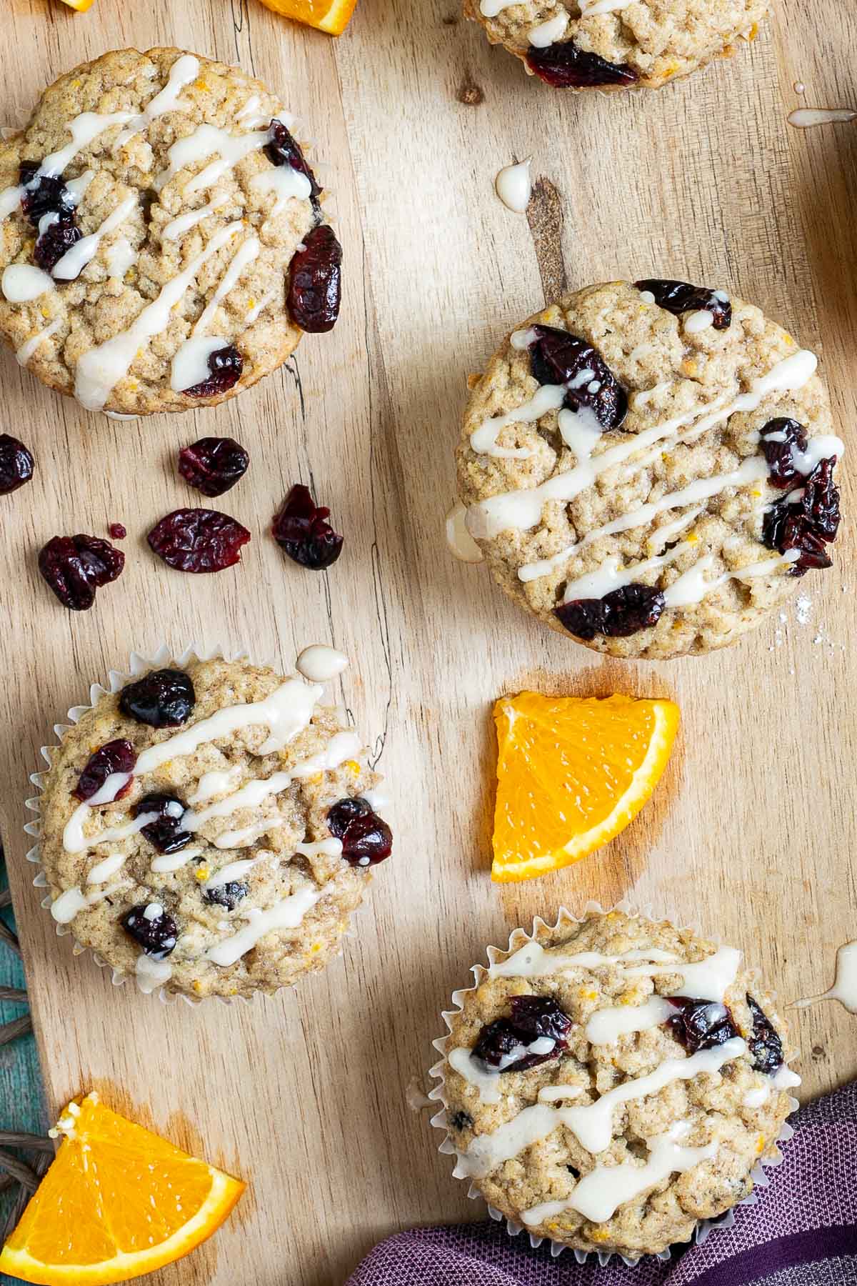Orange cranberry muffins drizzled with a white glaze on a wooden board surrounded by lots of dried cranberries and fresh orange slices.
