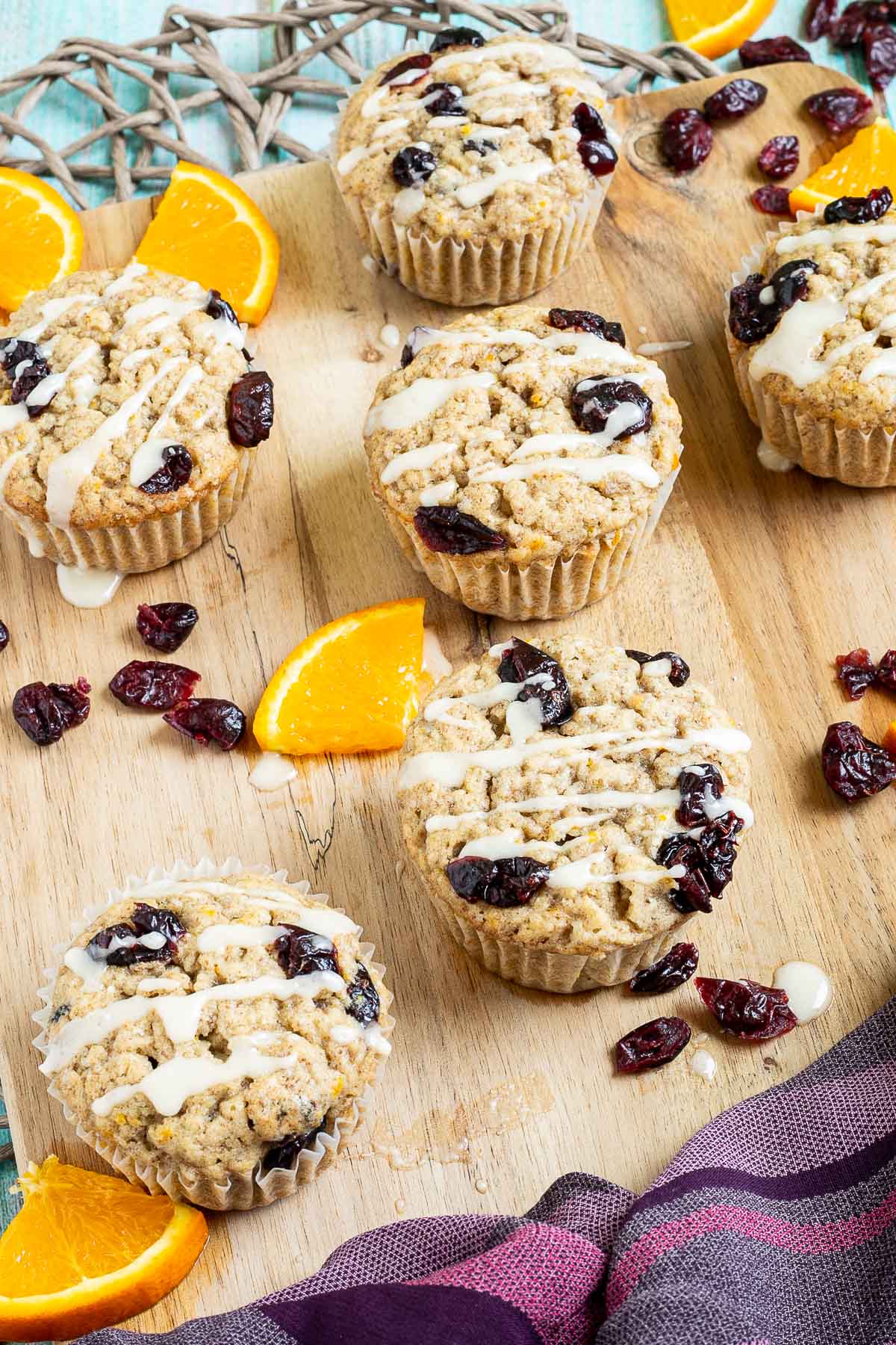 Orange cranberry muffins drizzled with a white glaze on a wooden board surrounded by lots of dried cranberries and fresh orange slices.