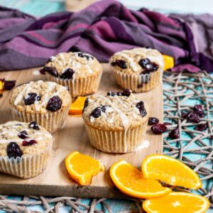 Orange cranberry muffins drizzled with a white glaze on a wooden board surrounded by lots of dried cranberries and fresh orange slices.