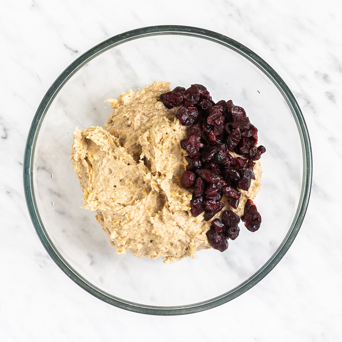 Glass bowl with light brown creamy batter topped with lots of dried cranberries.
