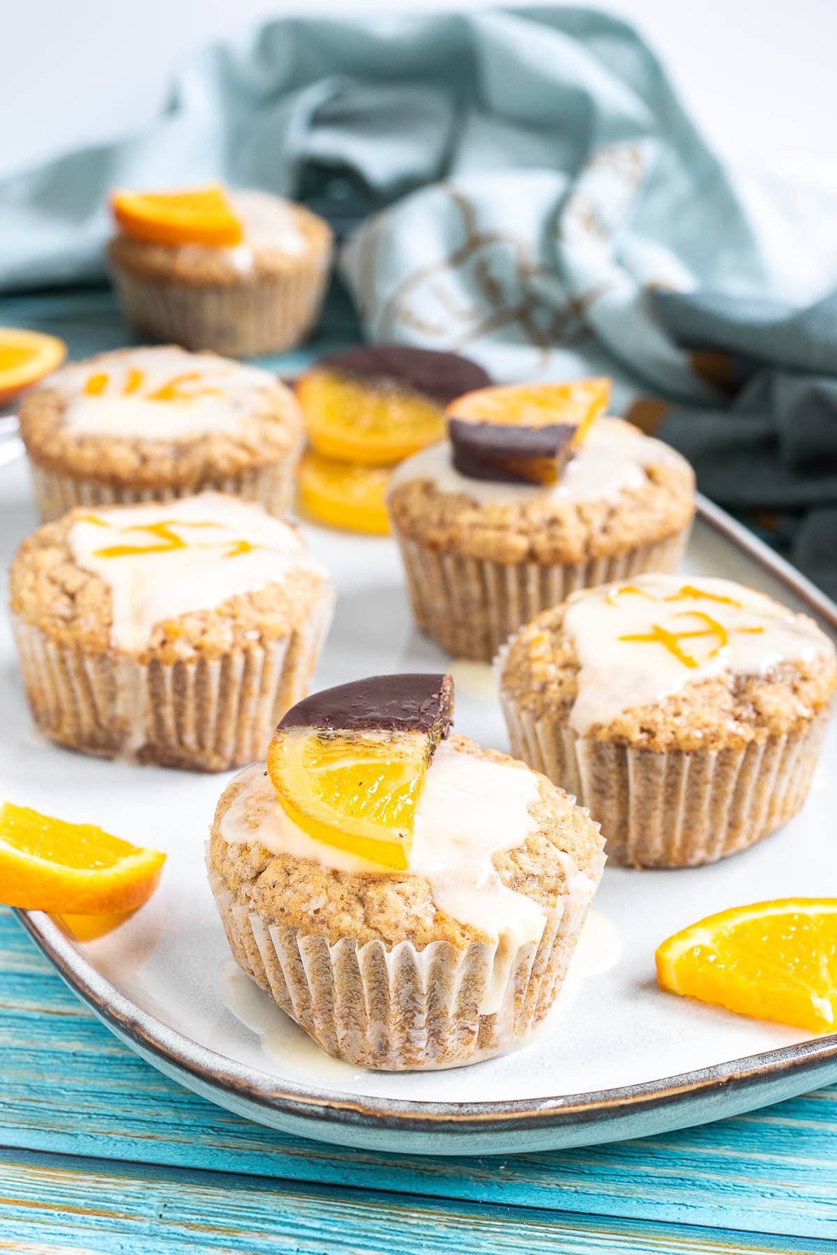 Several orange muffins on a light blue plate topped with a white glaze and orange slices partly dipped in chocolate or white glaze with orange zest pieces. Orange slices are scattered around them.