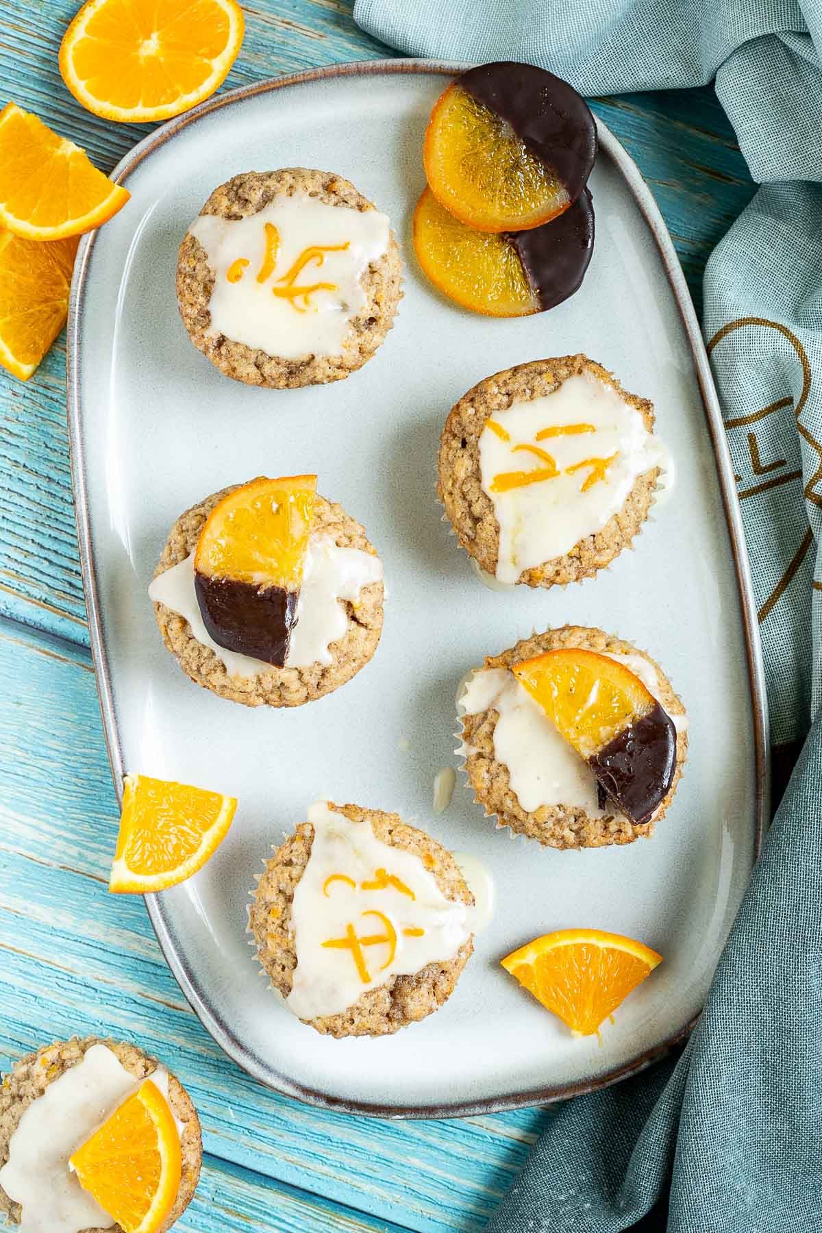 Several orange muffins on a light blue plate topped with a white glaze and orange slices partly dipped in chocolate or white glaze with orange zest pieces. Orange slices are scattered around them.