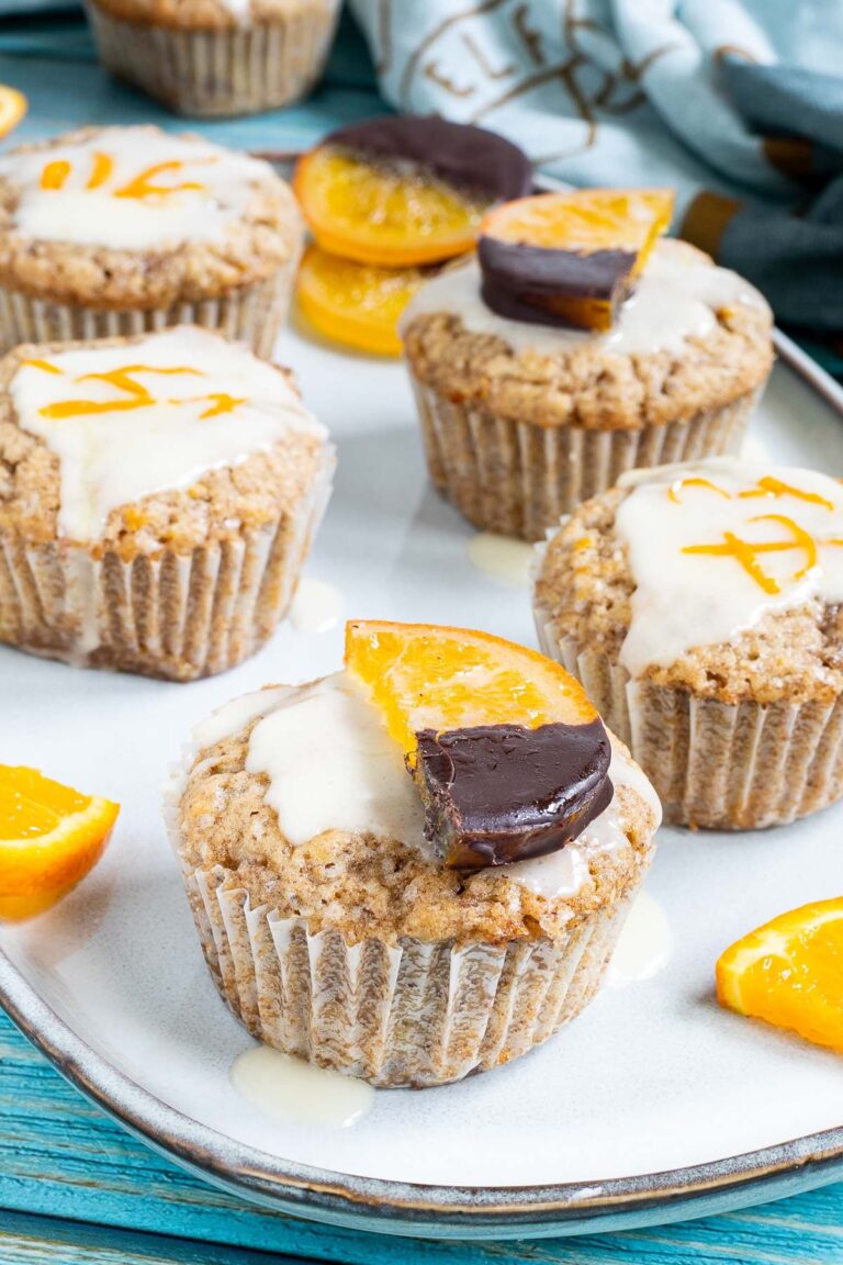 Several orange muffins on a light blue plate topped with a white glaze and orange slices partly dipped in chocolate or white glaze with orange zest pieces. Orange slices are scattered around them.