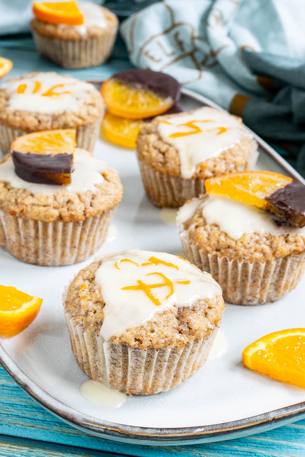 Several orange muffins on a light blue plate topped with a white glaze and orange slices partly dipped in chocolate or white glaze with orange zest pieces. Orange slices are scattered around them.