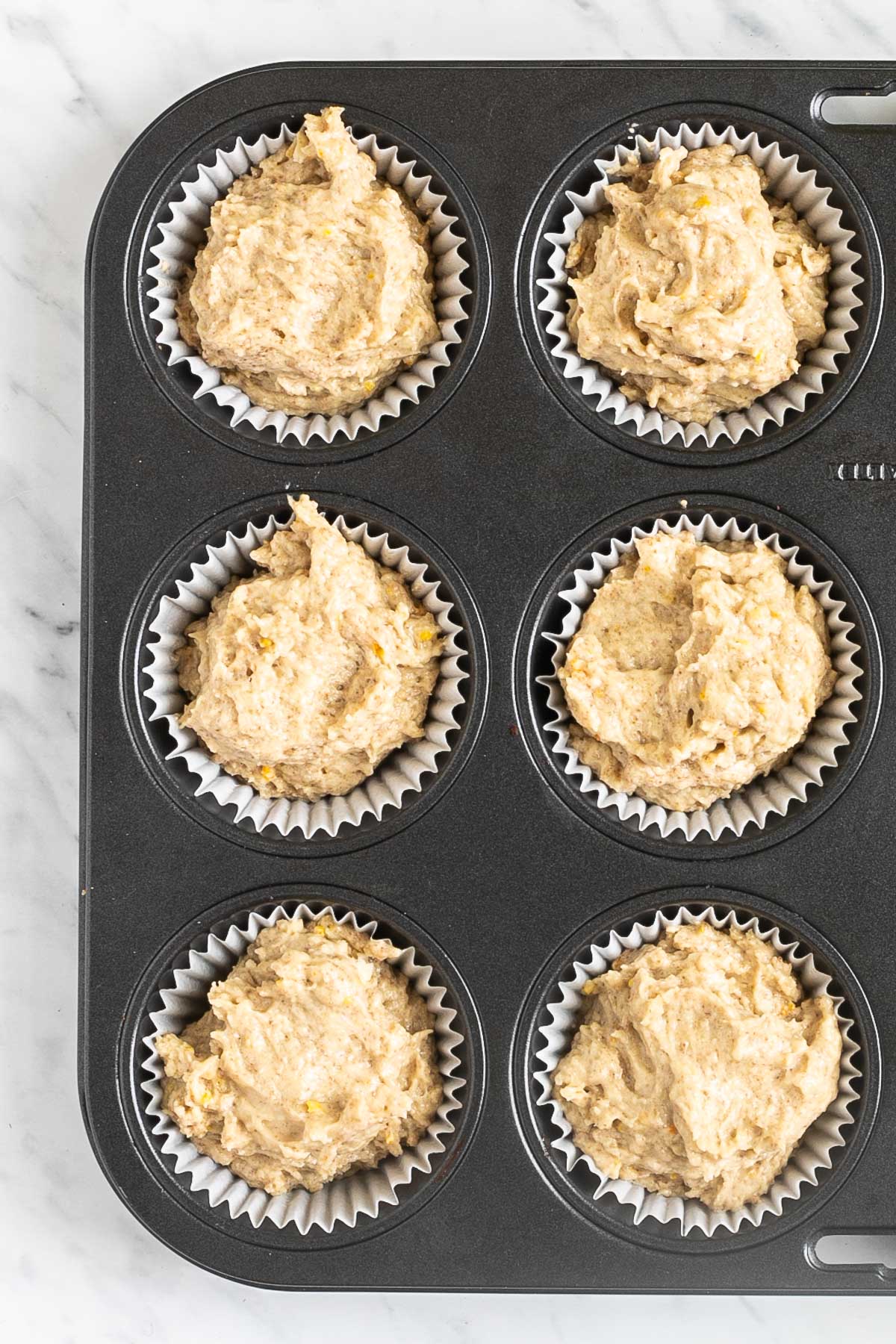 A muffin tin lined with white cupcake liners and filled with light brown batter.