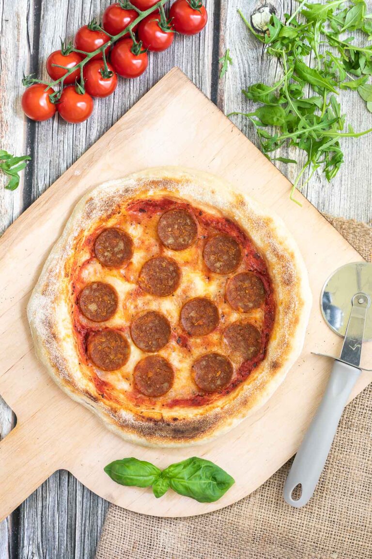 Pizza on a wooden baker's peel topped with tomato sauce, melted cheese and pepperoni slices. Fresh arugula, tomatoes and basil leaves are around it.