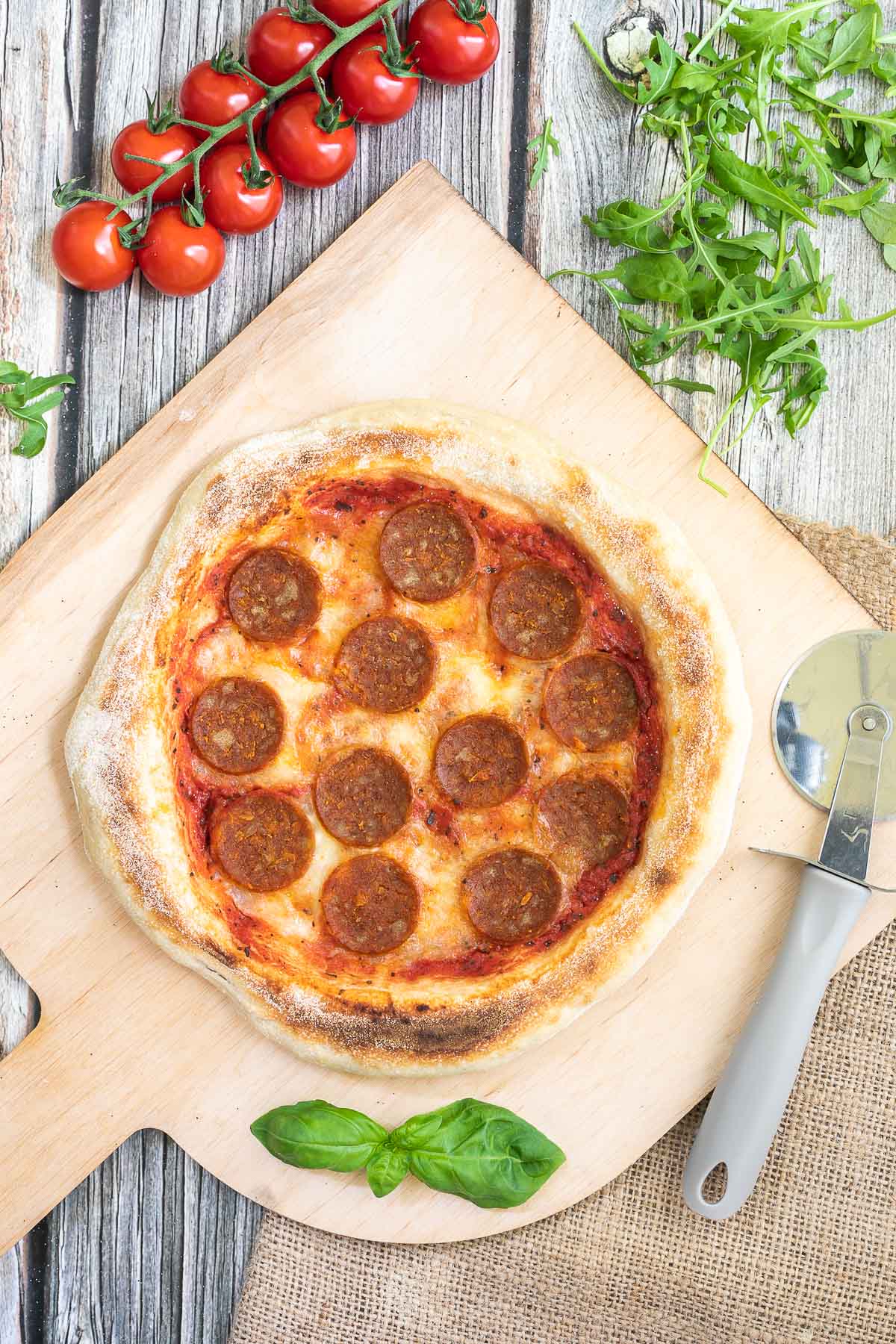 Pizza on a wooden baker's peel topped with tomato sauce, melted cheese and pepperoni slices. Fresh arugula, tomatoes and basil leaves are around it. 