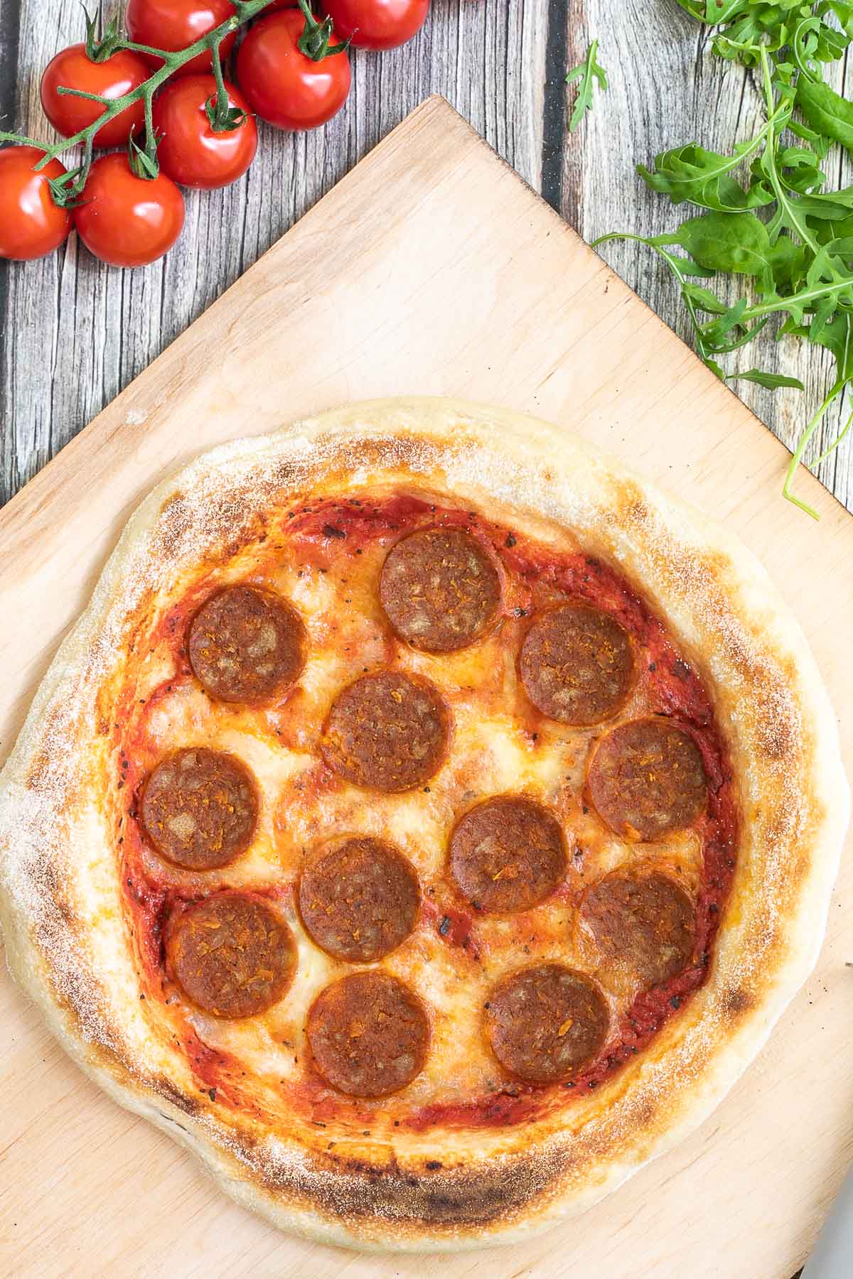Pizza on a wooden baker's peel topped with tomato sauce, melted cheese and pepperoni slices. Fresh arugula, tomatoes and basil leaves are around it.