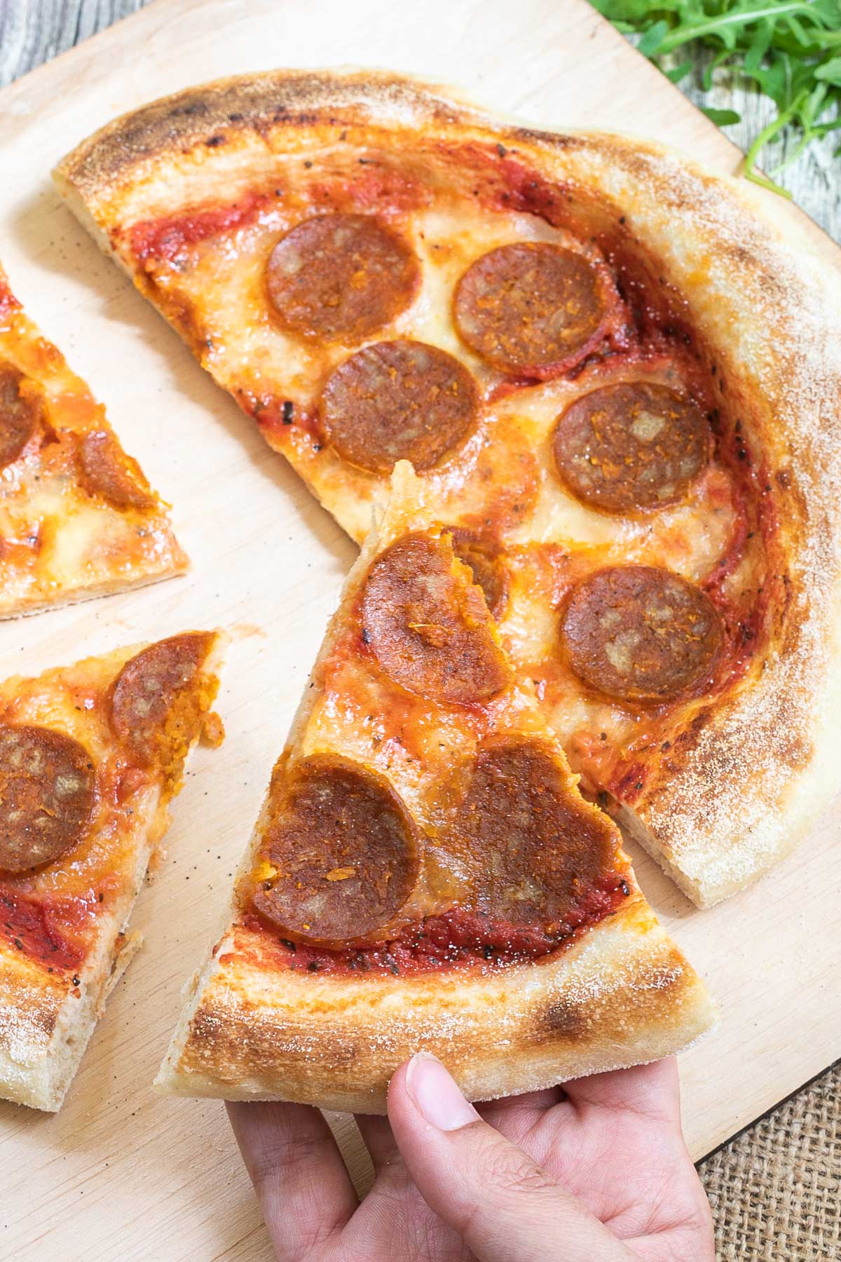 Sliced pizza on a wooden baker's peel topped with tomato sauce, melted cheese and pepperoni slices. A hand is taking a slice.