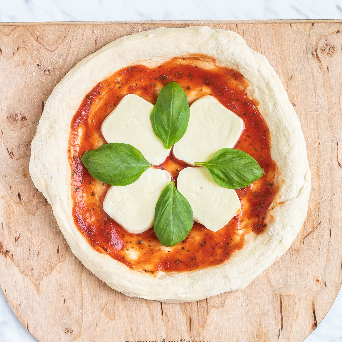 A pizza-shaped dough on a wooden pizza peel topped with tomato sauce, mozzarella and basil leaves.