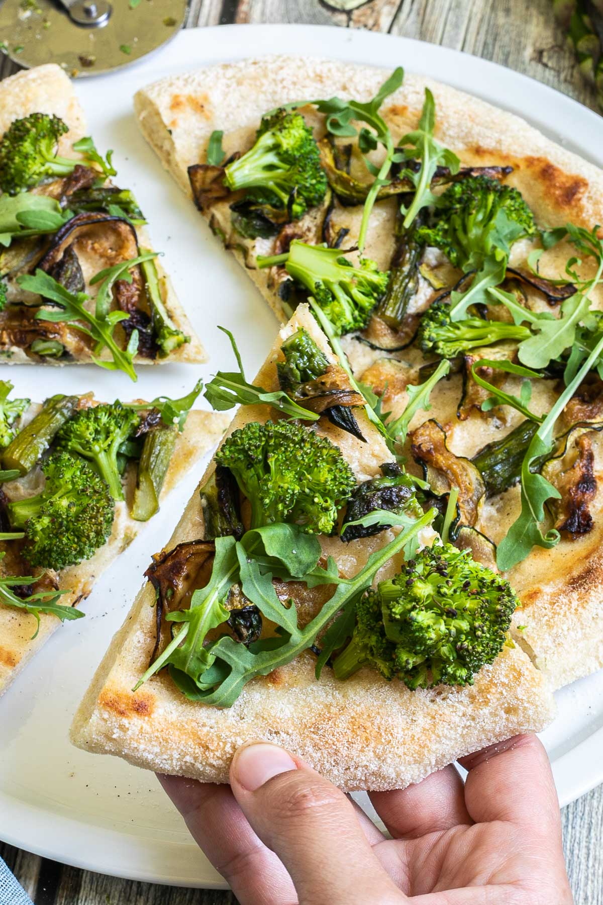 Sliced pizza on a white pizza plate topped with white sauce, zucchini, broccoli, asparagus, mushrooms, and fresh arugula. A hand is taking one slice.