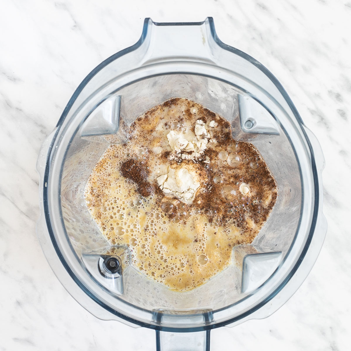 A blender from above with cashew nuts, milk, yellow flakes, flour, nutmeg, and salt.