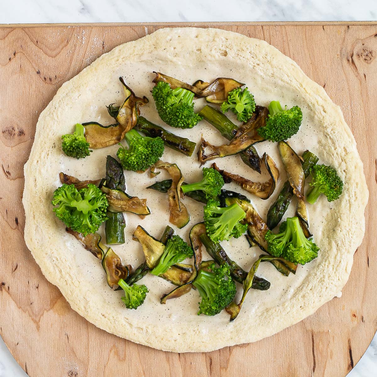 Unbaked pizza on a wooden baker's peel topped with white sauce, pan-fried zucchini, asparagus, and mushroom shreds, and broccoli.