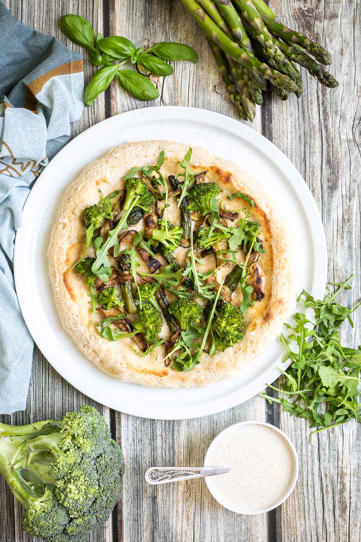 A pizza on a white pizza plate topped with white sauce, zucchini, broccoli, asparagus, mushrooms, and fresh arugula. More white sauce and fresh ingredients are scattered around the plate. 