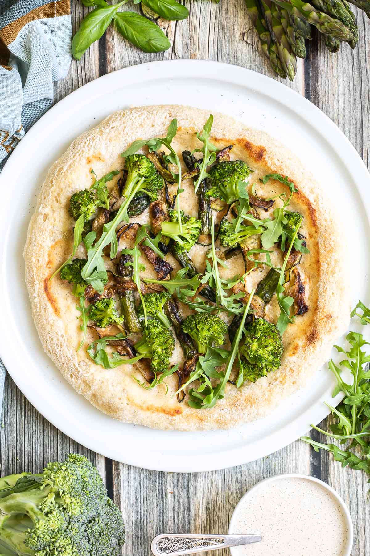 A pizza on a white pizza plate topped with white sauce, zucchini, broccoli, asparagus, mushrooms, and fresh arugula. More white sauce and fresh ingredients are scattered around the plate.