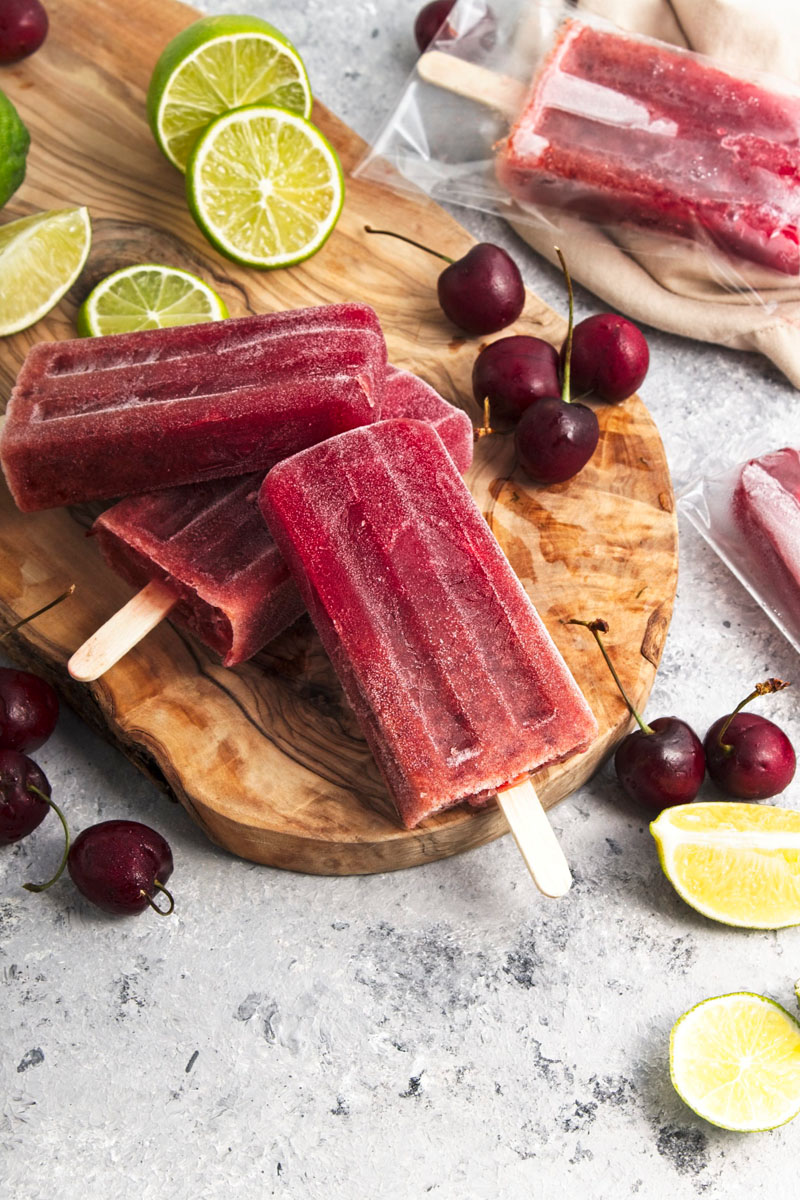 Vibrant purple popsicles on a wooden board. Cherries and lime slices are around them.