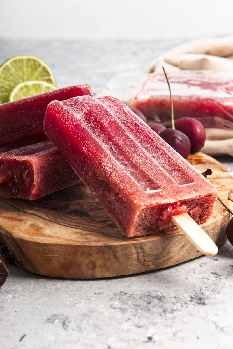 Vibrant purple popsicles on a wooden board. Cherries and lime slices are around them.