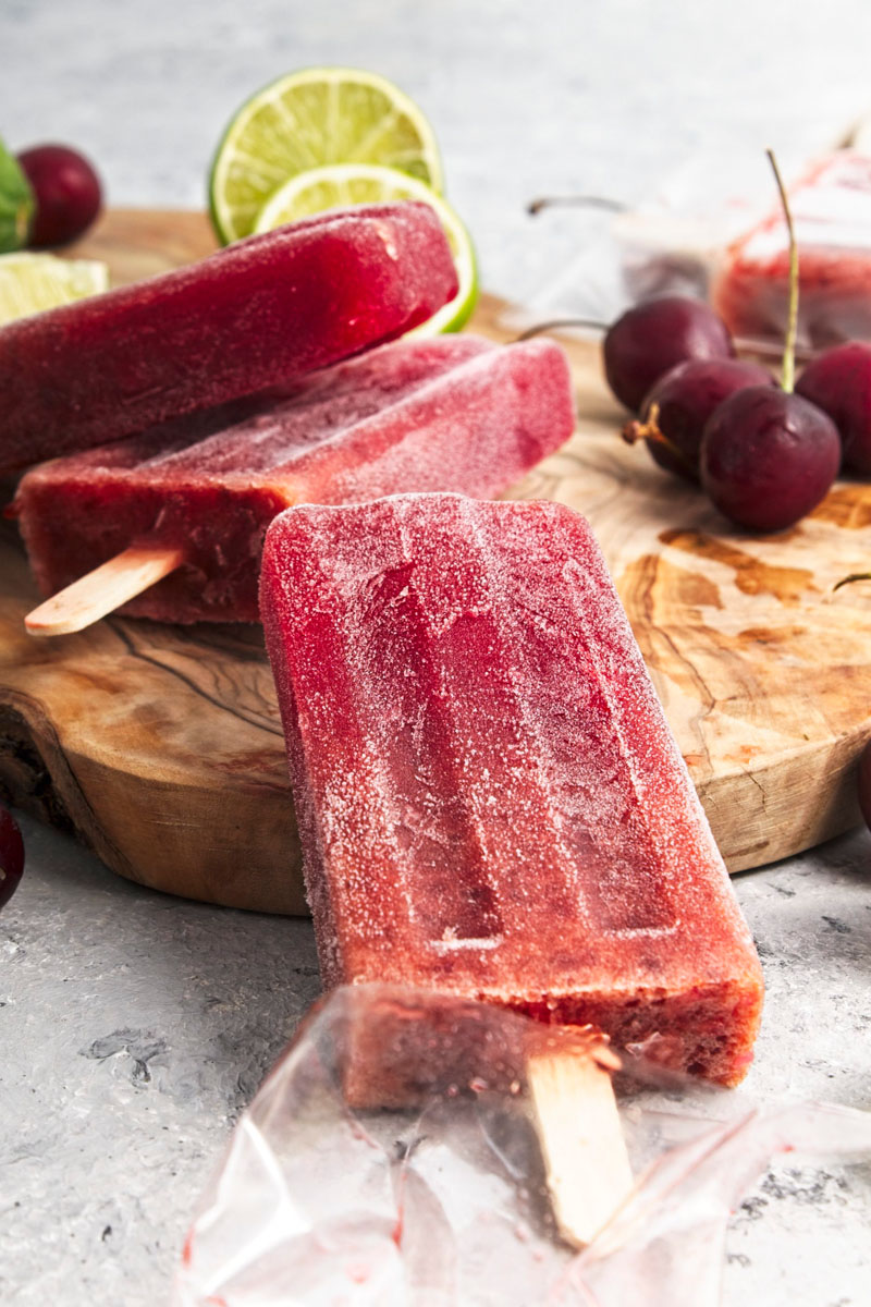 Vibrant purple popsicles on a wooden board. Cherries and lime slices are around them.