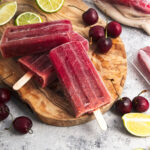 Vibrant purple popsicles on a wooden board. Cherries and lime slices are around them.