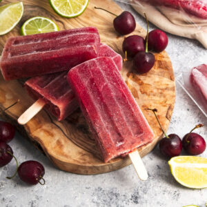 Vibrant purple popsicles on a wooden board. Cherries and lime slices are around them.
