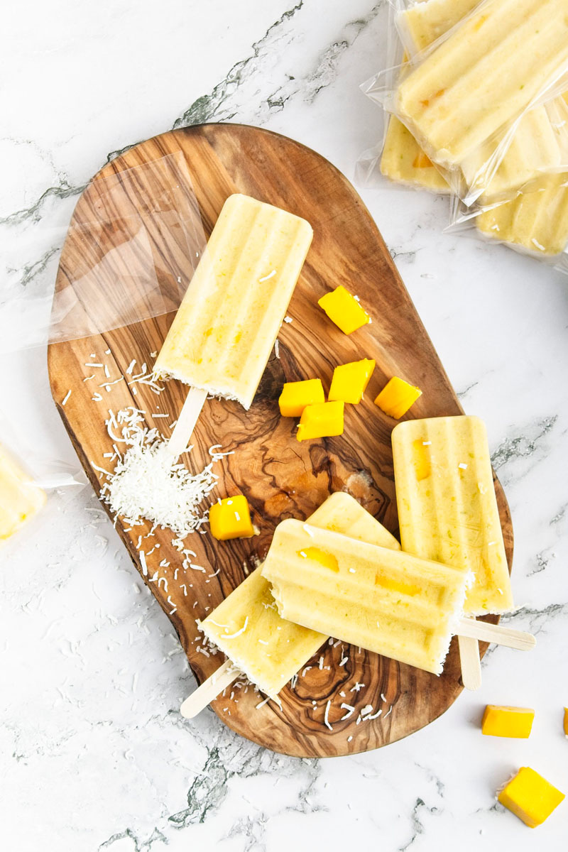 Vibrant yellow popsicles on a wooden board. Mango pieces and shredded coconut is around them.