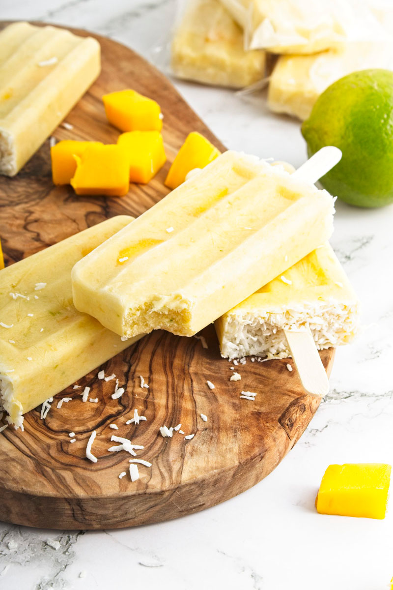 Vibrant yellow popsicles on a wooden board. Mango pieces and shredded coconut is around them. A bit is missing from one of them.