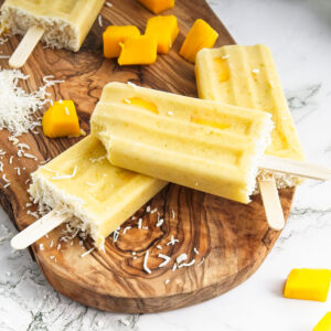 Vibrant yellow popsicles on a wooden board. Mango pieces and shredded coconut is around them. A bit is missing from one of them.