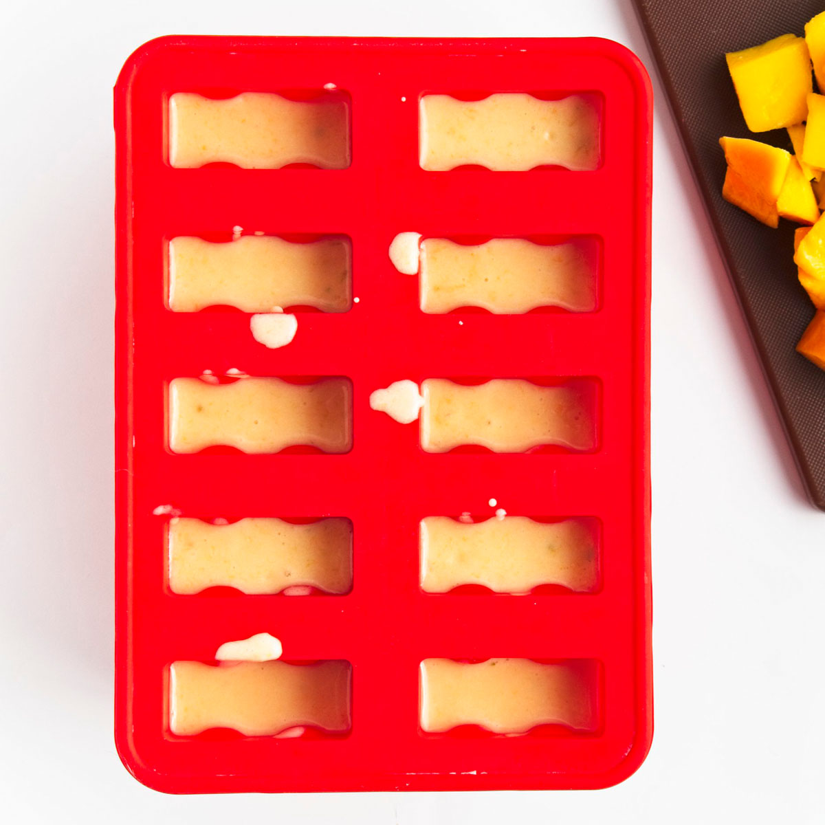 Red popsicle mold with 10 holes filled to the brim with yellow puree.