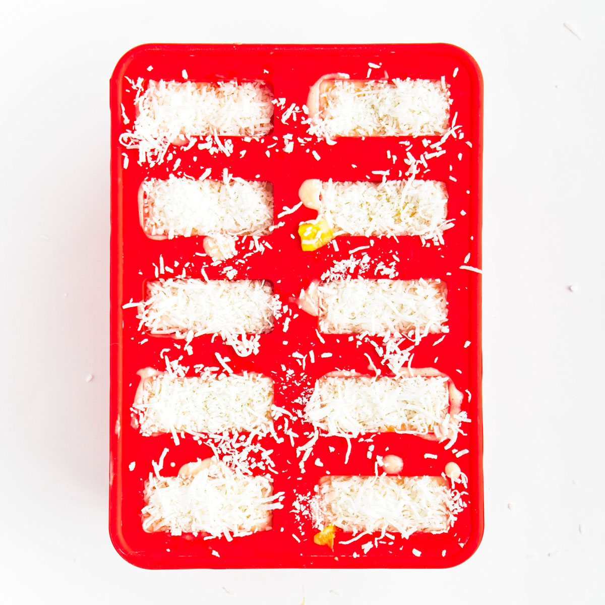 Red popsicle mold with 10 holes filled to the brim with yellow puree topped with shredded coconut.