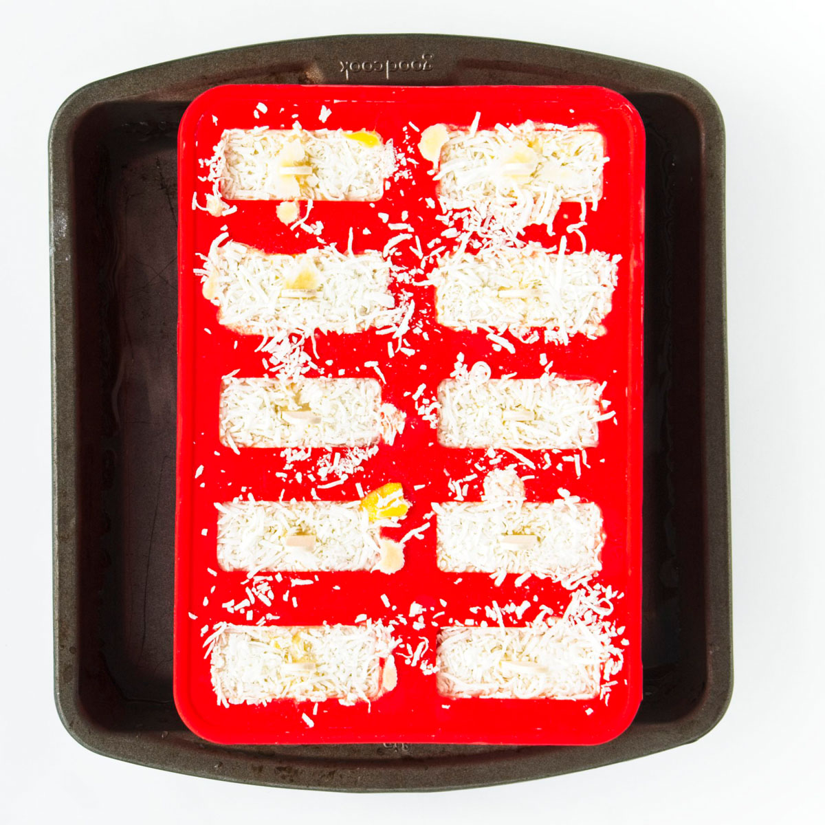 Red popsicle mold with 10 holes filled to the brim with sticks sticking out from them in a large black pan filled with water.