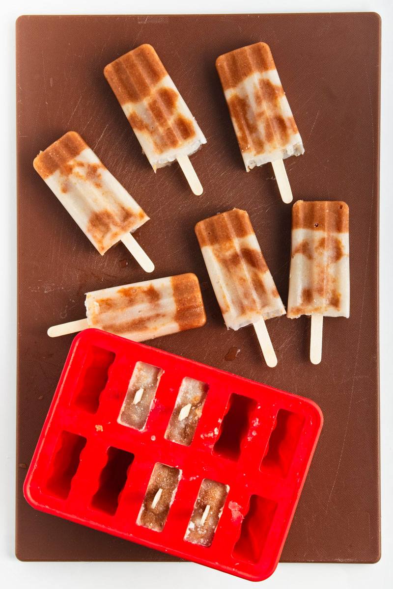 Brown and white popsicles on a brown board after they were unmolded from a red popsicle mold placed next to them.
