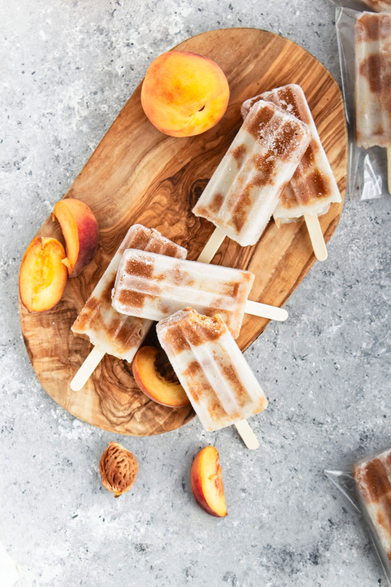 Brown and white popsicles on a wooden cutting board with peach slices scattered around them.