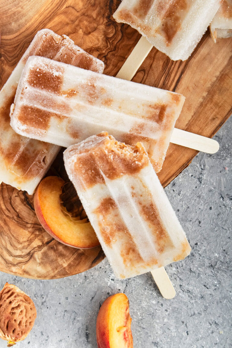 Brown and white popsicles on a wooden cutting board with peach slices scattered around them.