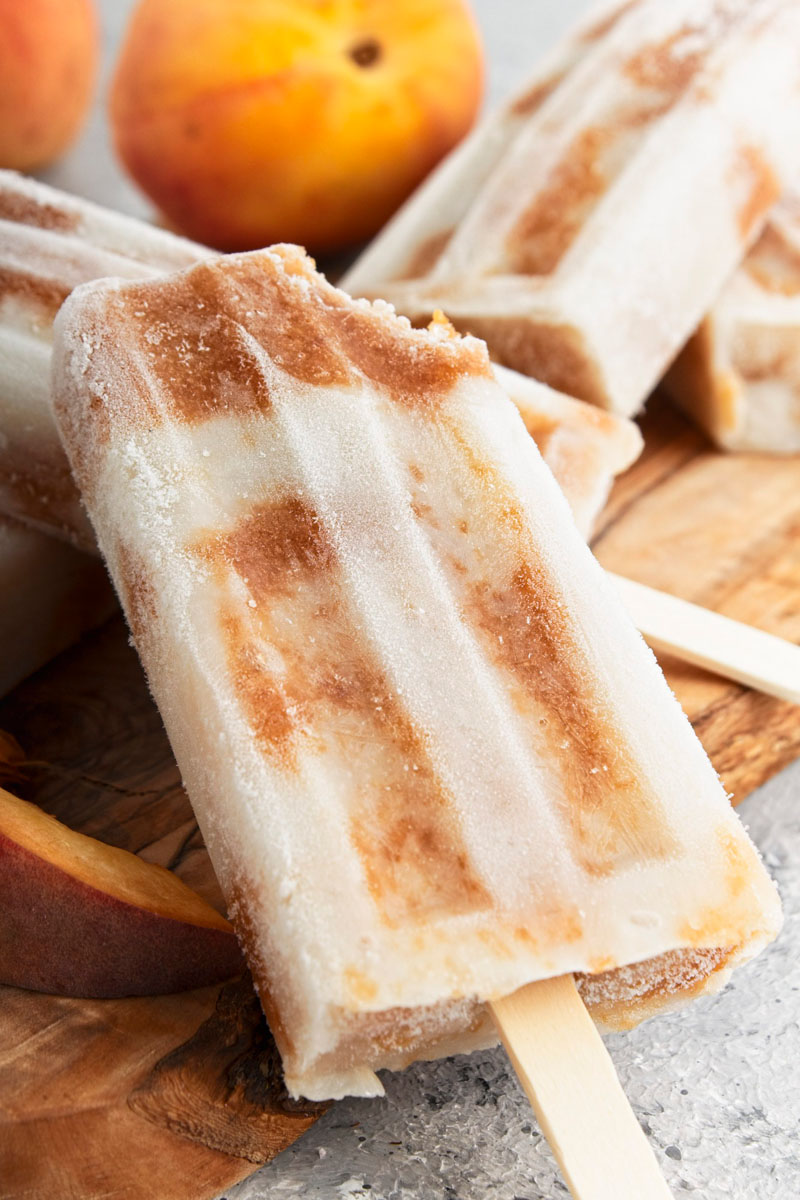 Brown and white popsicles on a wooden cutting board with whole peach at the background.