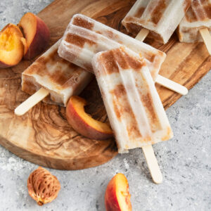 Brown and white popsicles on a wooden cutting board with peach slices scattered around them.
