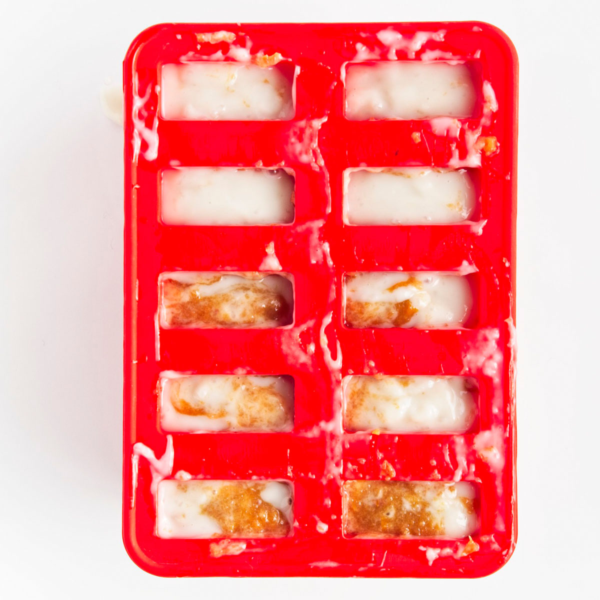 Red popsicle mold with 10 holes filled to the brim with brown and white puree.