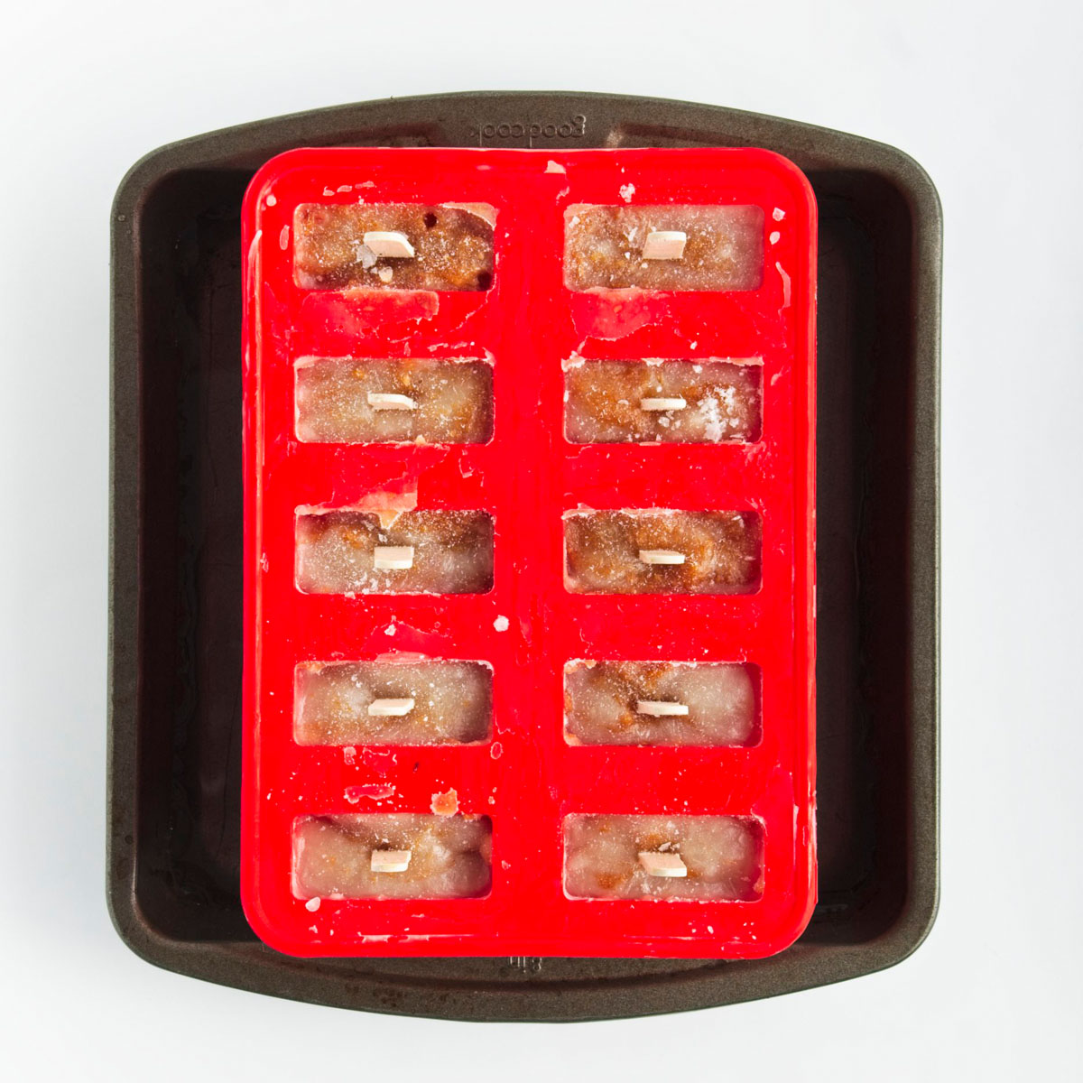 Red popsicle mold with 10 holes filled to the brim with sticks sticking out from them in a large black pan filled with water.