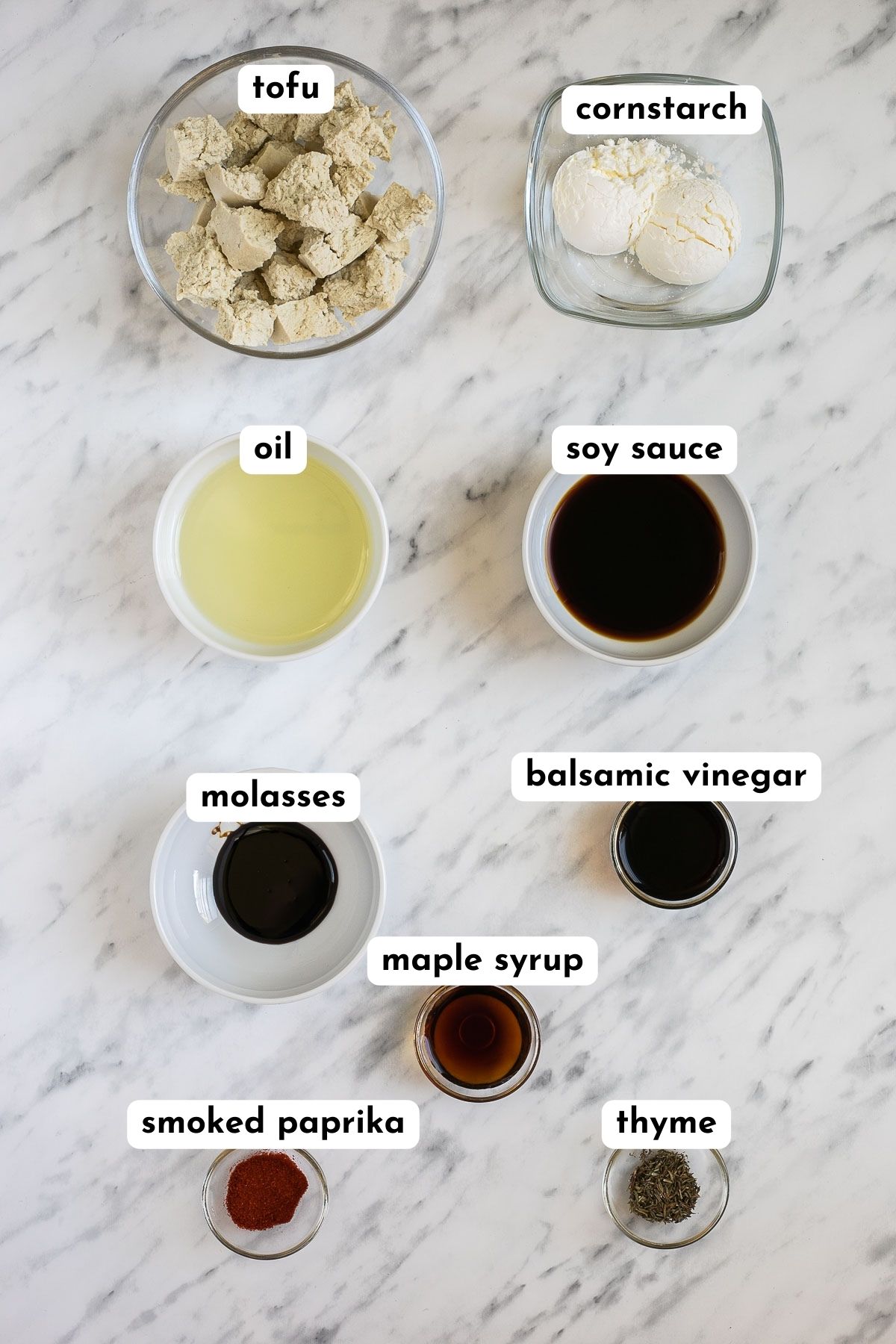 Ingredients of marinated tofu in small glass bowls like tofu pieces, soy sauce, molasses, balsamic vinegar, starch, oil, maple syrup, thyme and paprika powder