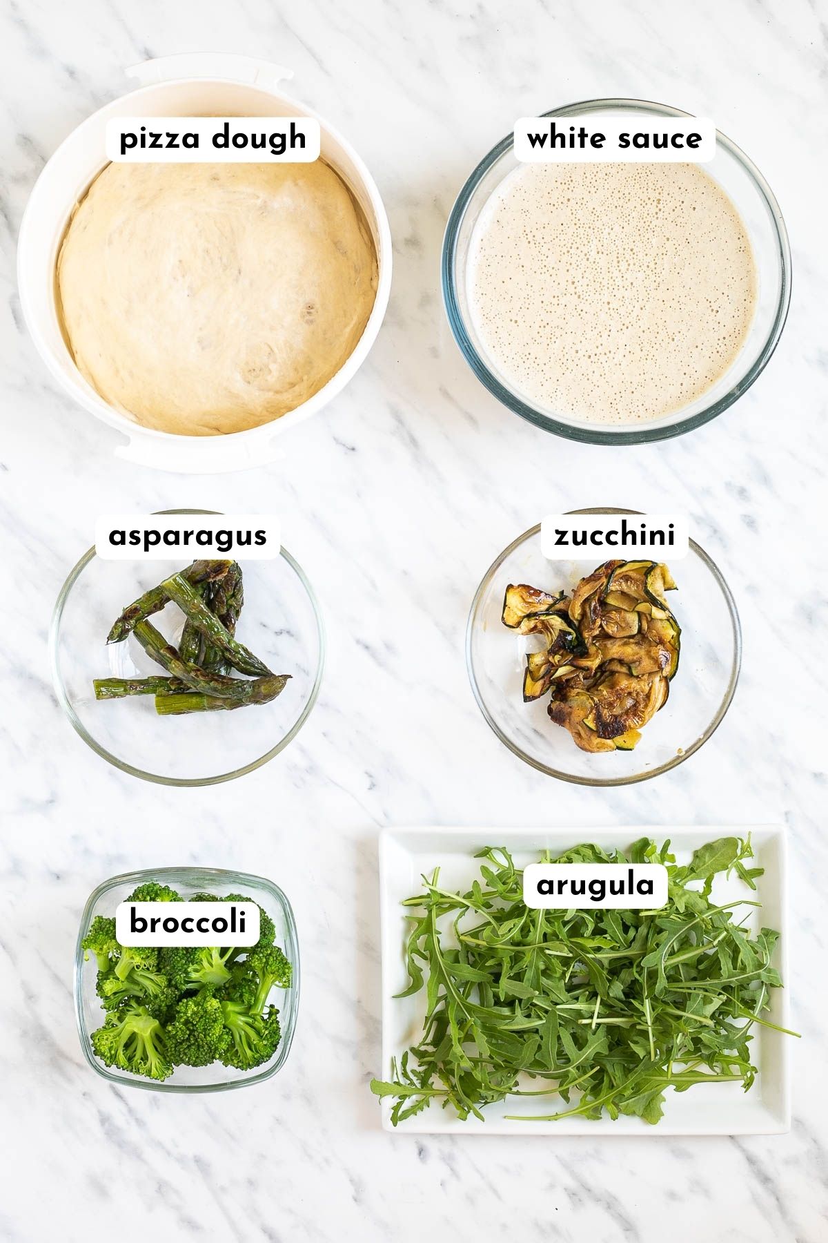 Ingredients of vegan white pizza in small containers like pizza dough, white sauce, asparagus, zucchini, broccoli, arugula