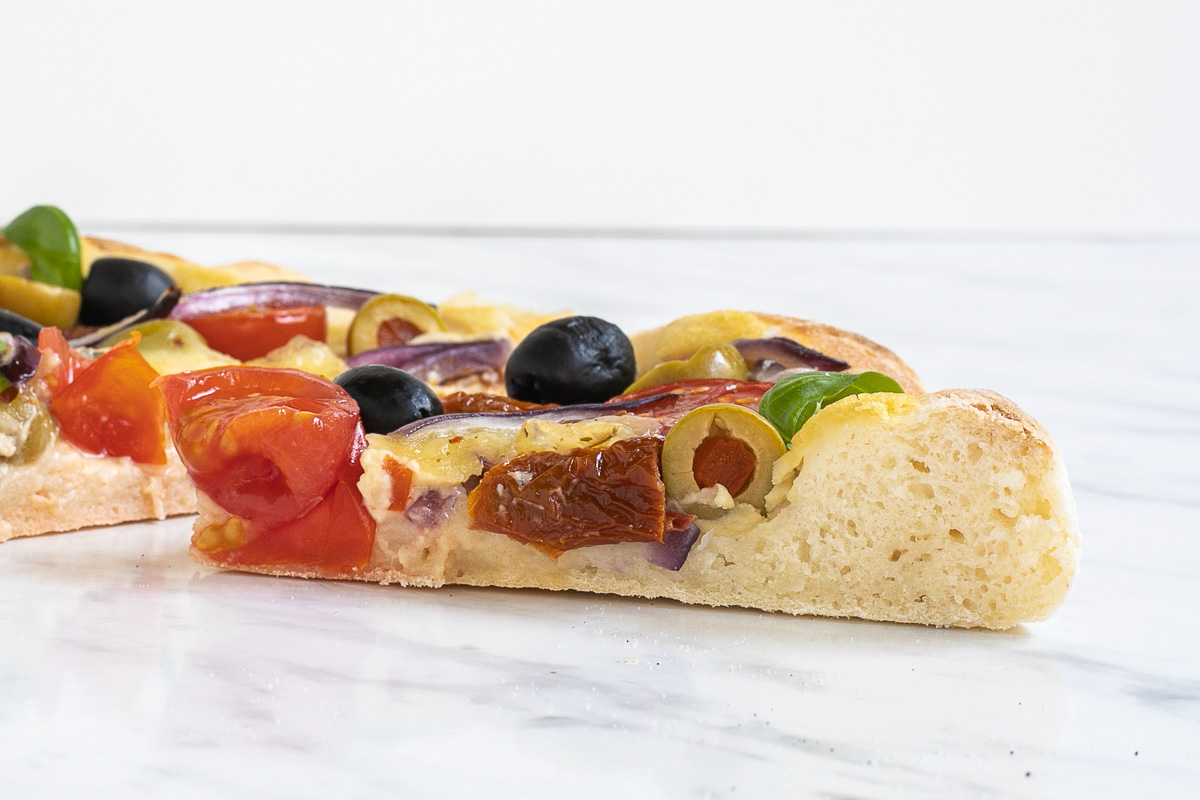 A slice of pizza with olives, cherry tomatoes, red onion slices as topping from the side to see the airy fluffy rim.