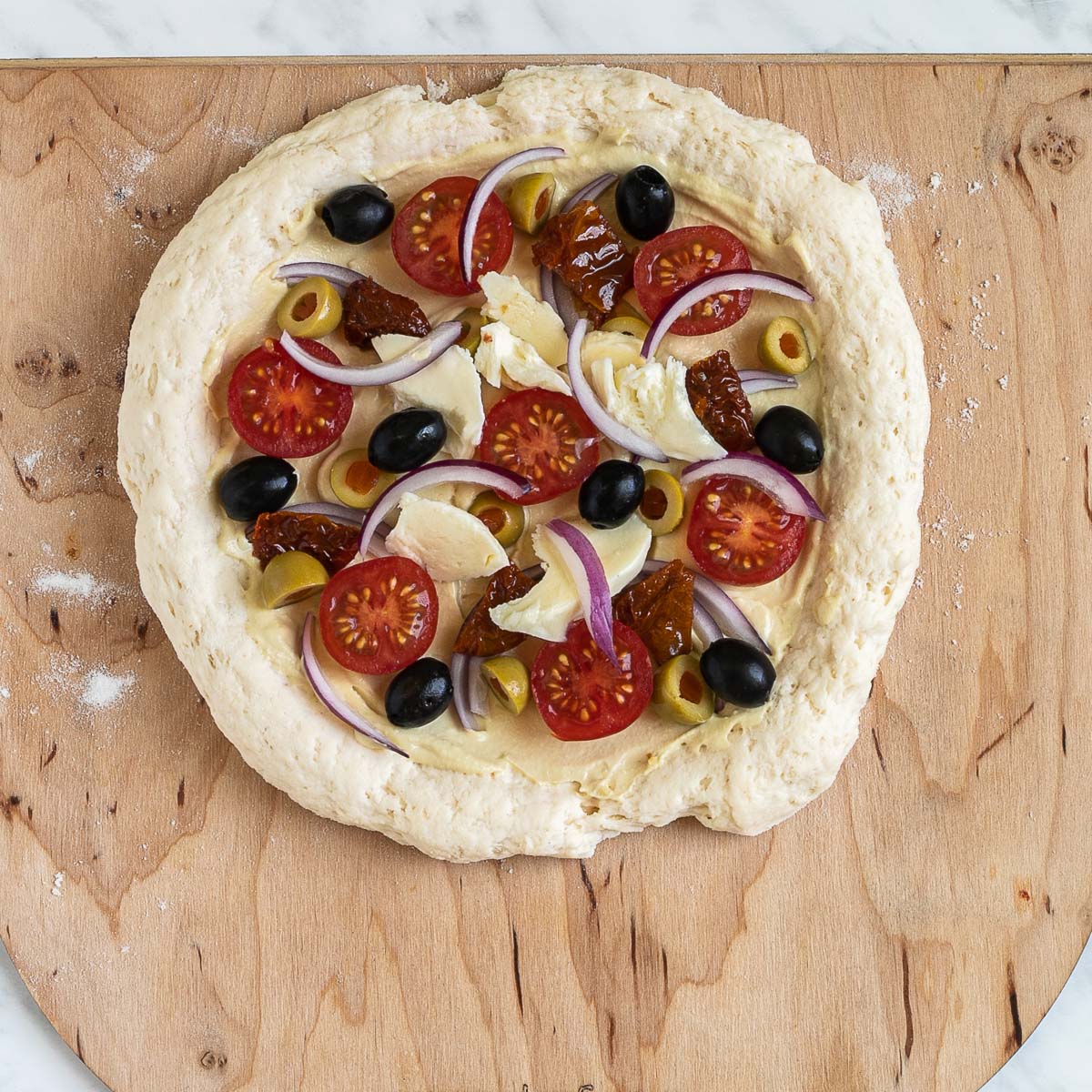 Unbaked pizza on a wooden baker's peel topped with tomatoes, green and black olives, cheese, red onion slices.