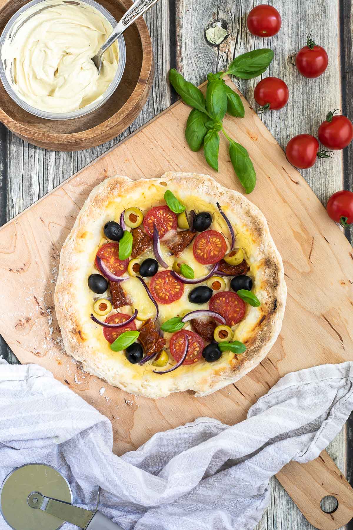 Pizza on a wooden baker's peel topped with light brown hummus, cherry tomatoes, green and black olives, red onion slices, and fresh basil leaves. More hummus and other fresh ingredients are scattered around it.