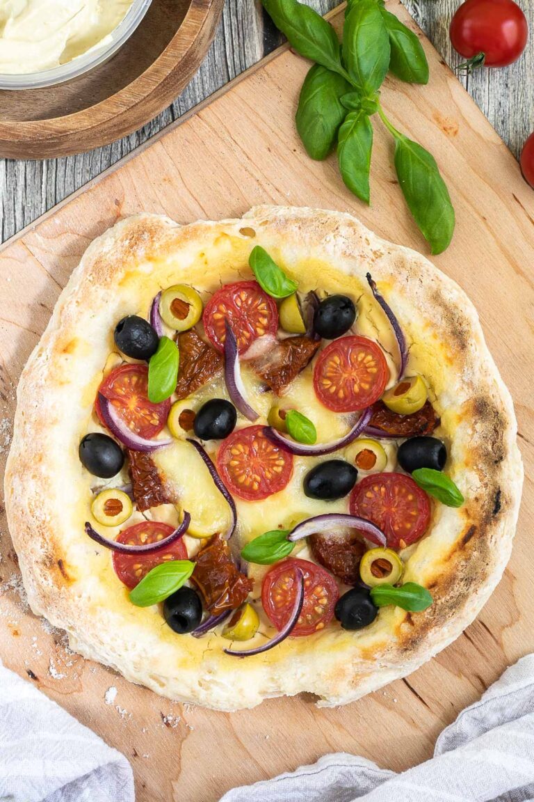 Pizza on a wooden baker's peel topped with light brown hummus, cherry tomatoes, green and black olives, red onion slices, and fresh basil leaves. More hummus and other fresh ingredients are scattered around it.