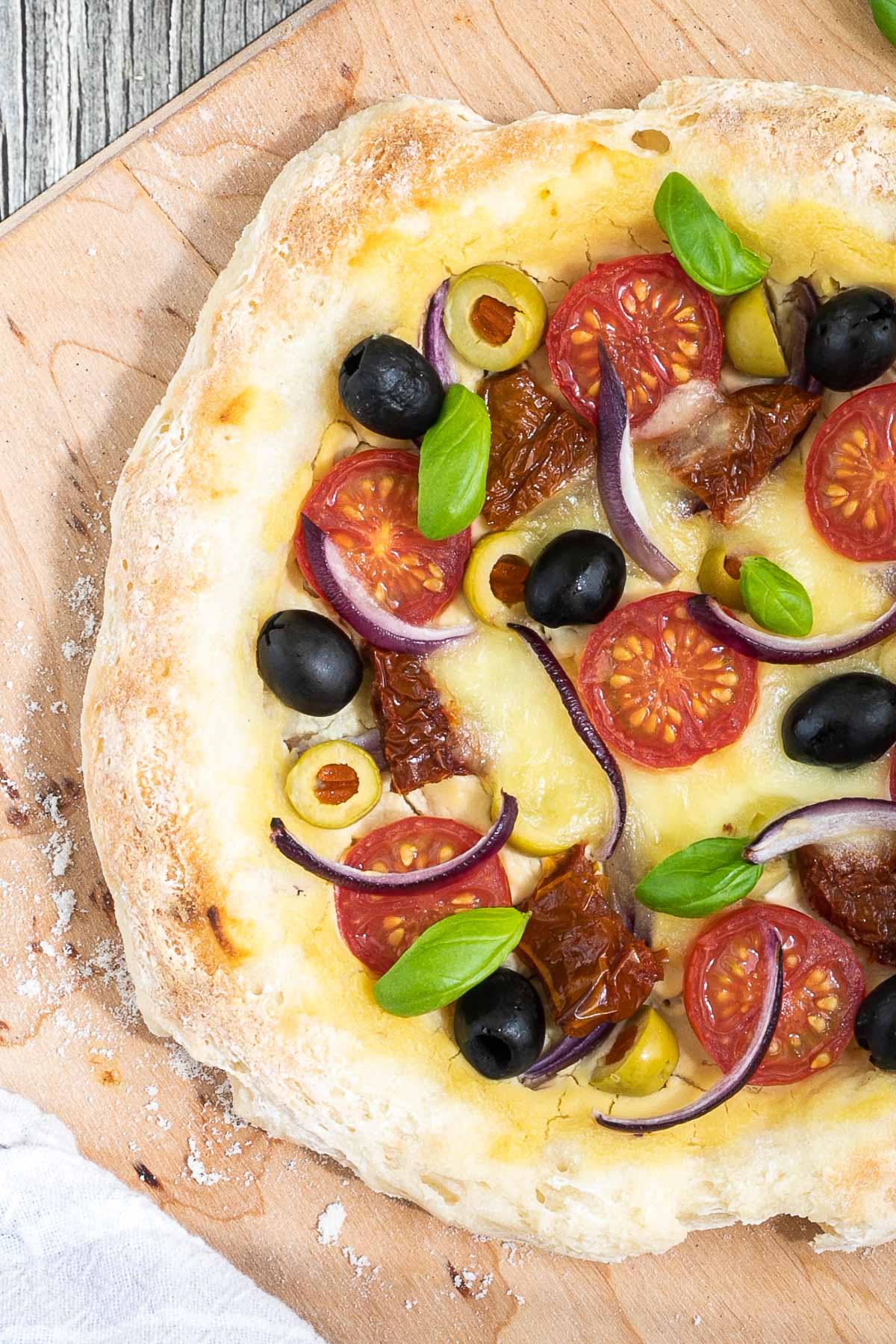 Pizza on a wooden baker's peel topped with light brown hummus, cherry tomatoes, green and black olives, red onion slices, and fresh basil leaves.