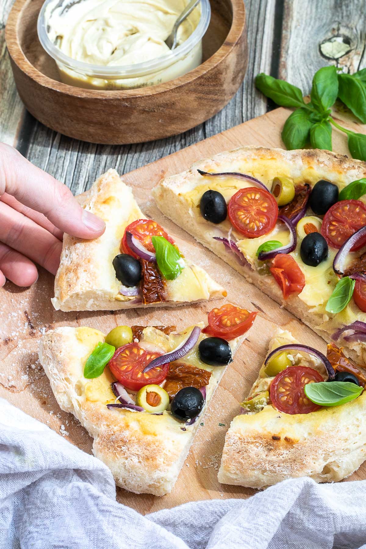 Sliced pizza on a wooden baker's peel topped with light brown hummus, cherry tomatoes, green and black olives, red onion slices, and fresh basil leaves. A hand is talking a slice. More hummus and other fresh ingredients are scattered around it.