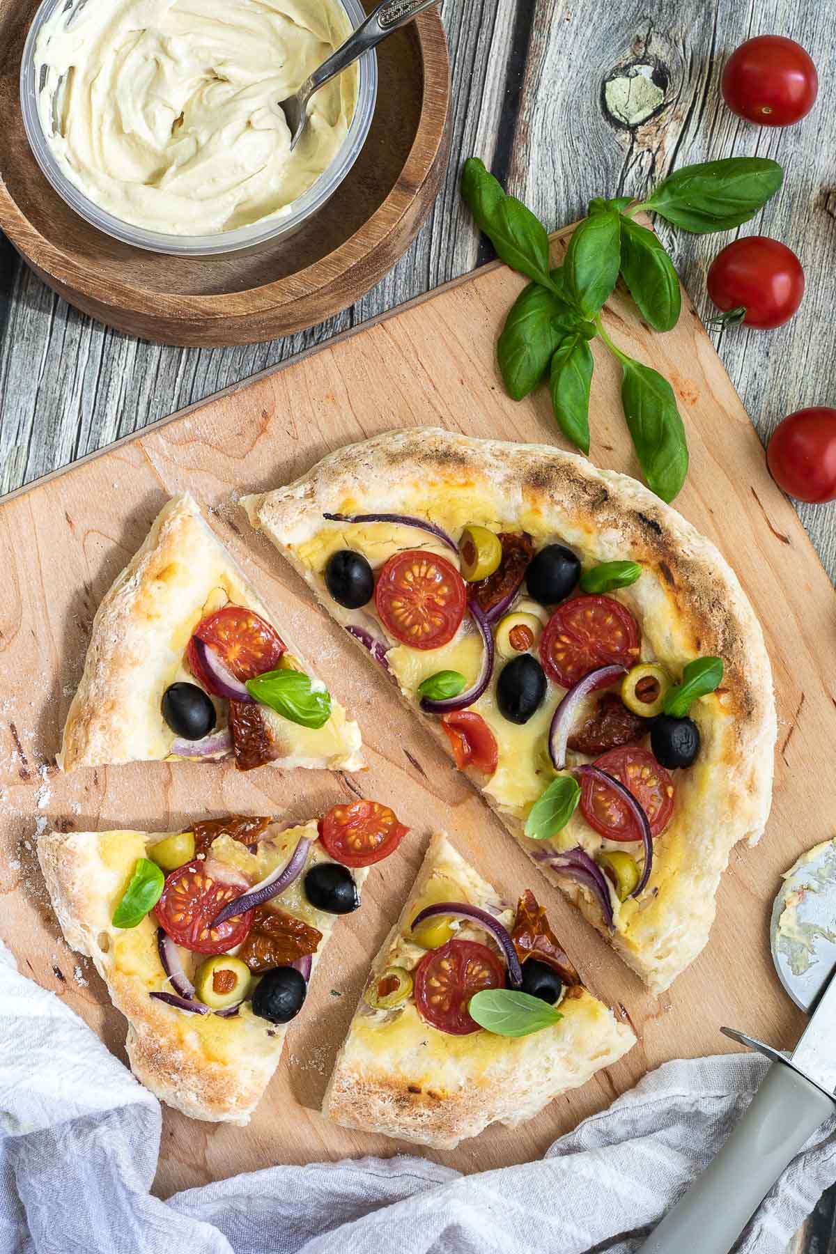 Sliced pizza on a wooden baker's peel topped with light brown hummus, cherry tomatoes, green and black olives, red onion slices, and fresh basil leaves. More hummus and other fresh ingredients are scattered around it.