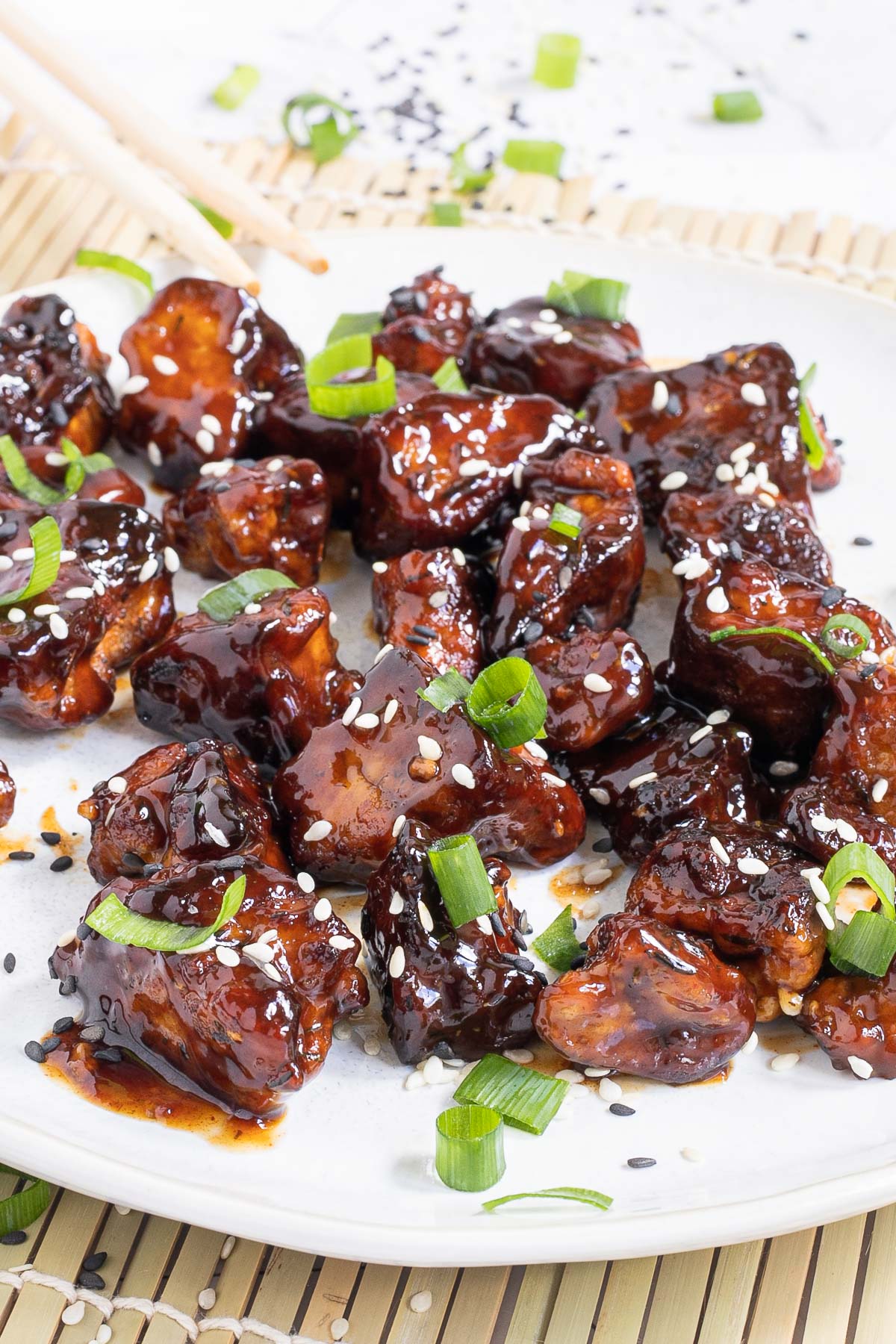 Dark brown sticky juice tofu pieces on a white plate sprinkled with black and white sesame seeds and chopped scallion.