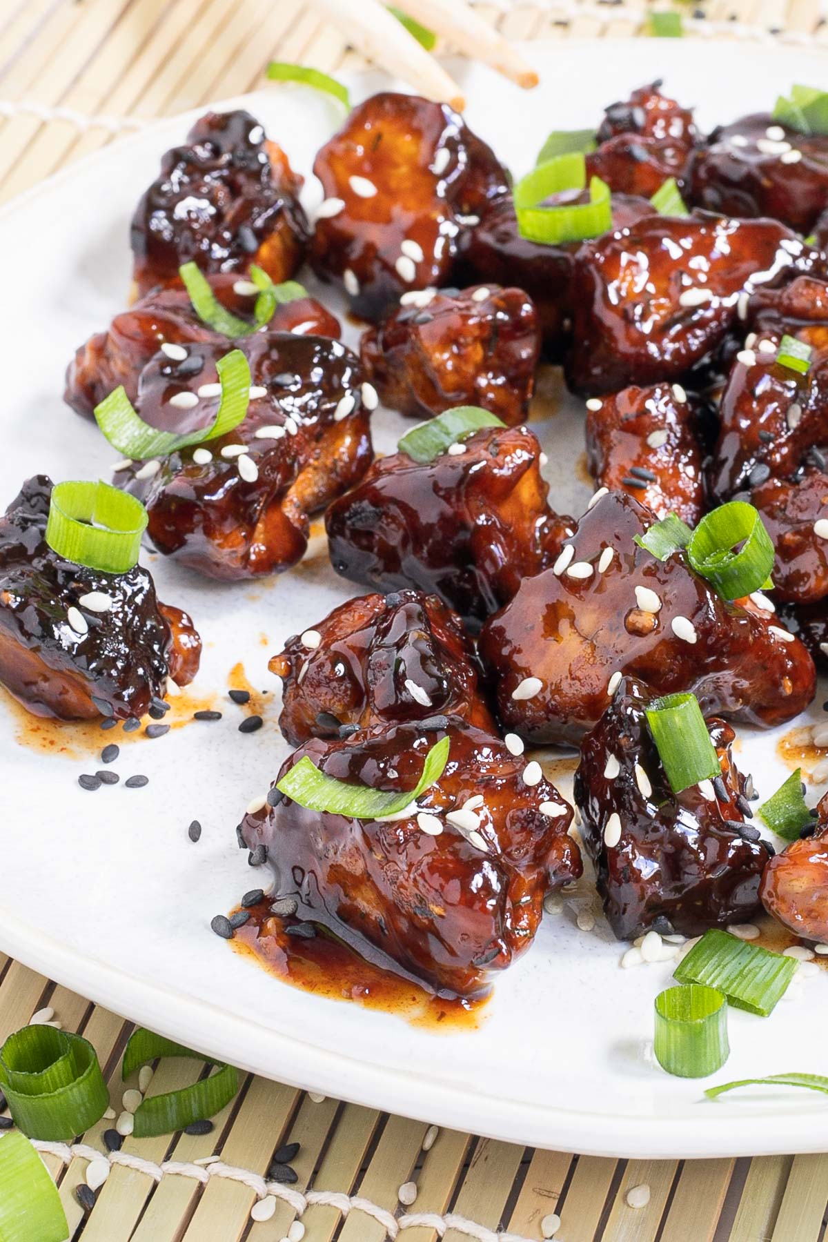 Dark brown sticky juice tofu pieces on a white plate sprinkled with black and white sesame seeds and chopped scallion.
